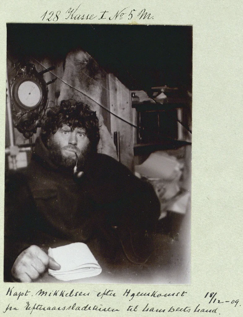 Portrait of Mikkelsen after his sledding trip, with long hair and beard, smoking a pipe